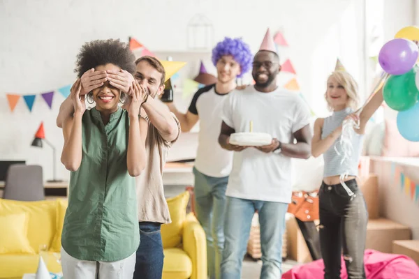 Young men and women covering eyes of young female friend and greeting her with birthday cake on surprise party — Stock Photo