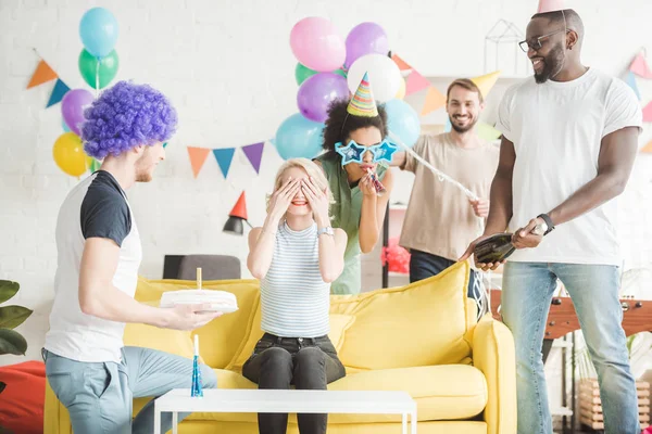 Lächelnde junge Leute begrüßen junge Frau mit Geburtstagstorte auf Überraschungsparty — Stockfoto