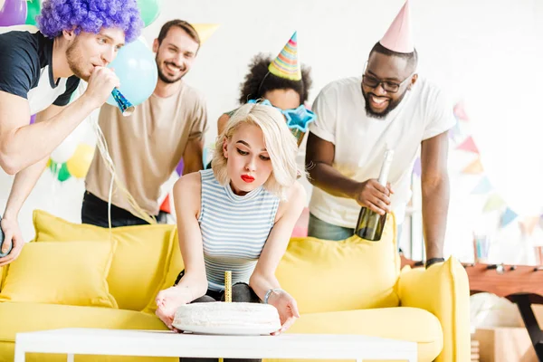 Chica feliz soplando vela en pastel de cumpleaños por sus amigos alegres - foto de stock