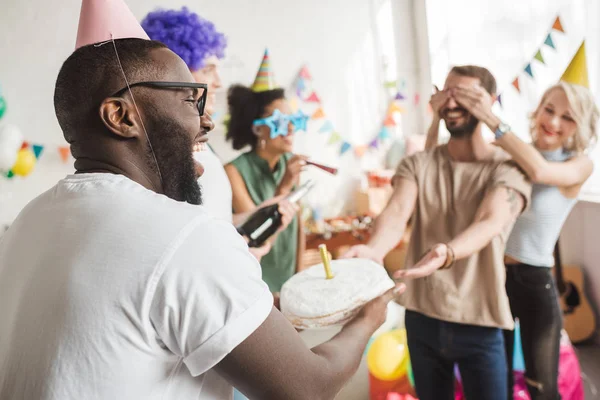 Щасливі друзі прикривають очі молодого чоловіка і вітають його з тортом на день народження — стокове фото