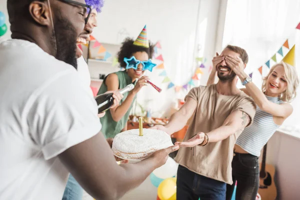 Partying diverse people covering eyes of young friend and greeting him with birthday cake — Stock Photo