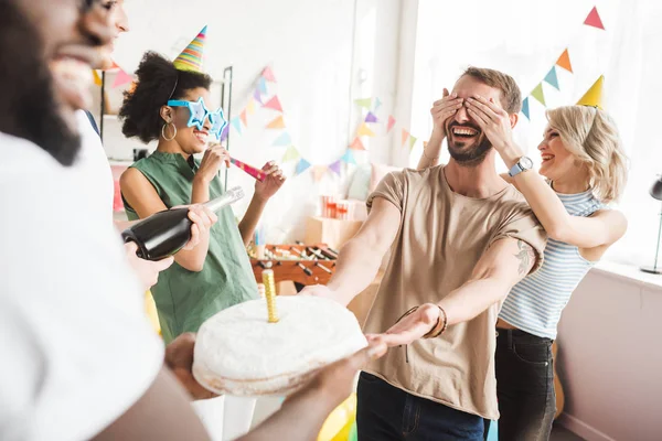 Lächelnde junge Leute bedecken die Augen eines jungen Freundes und grüßen ihn mit Geburtstagstorte — Stockfoto