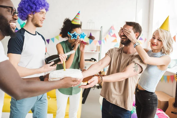 Young men and women covering eyes of young friend and greeting him with birthday cake — Stock Photo