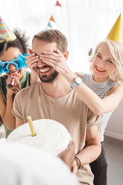 Young people covering eyes of young man and greeting him with birthday cake — Stock Photo