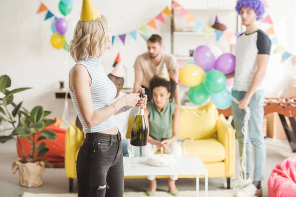 Blonde girl opening champagne in front of partying friends — Stock Photo