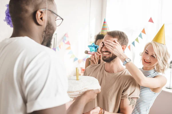 Glückliche Freunde bedecken die Augen des jungen Mannes und begrüßen ihn mit Geburtstagstorte — Stockfoto