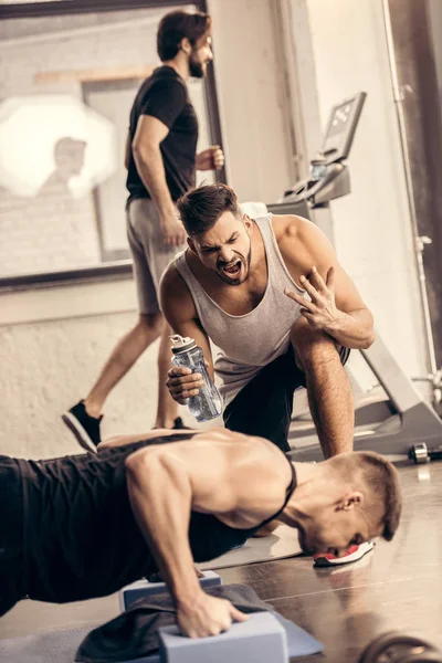 Trainer schreit Sportler beim Planken auf Yoga-Blöcken im Fitnessstudio an — Stockfoto