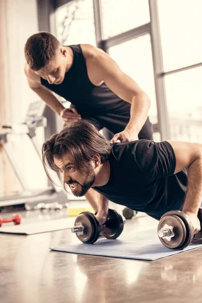 Entrenador empujando deportista haciendo flexiones en mancuernas en el gimnasio - foto de stock