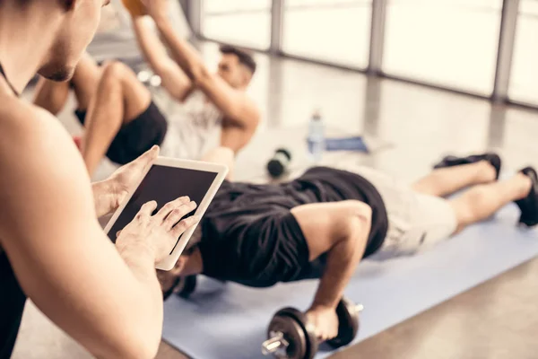 Treinador usando tablet e contando resultados de desportista fazendo flexões em halteres no ginásio — Fotografia de Stock