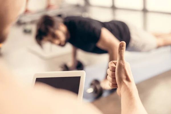 Trainer holding tablet and showing thumb up to sportsman doing push ups on dumbbells in gym — Stock Photo