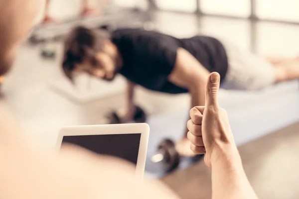 Entraîneur tenant la tablette et montrant pouce jusqu'à sportif faisant des pompes sur haltères dans la salle de gym — Photo de stock