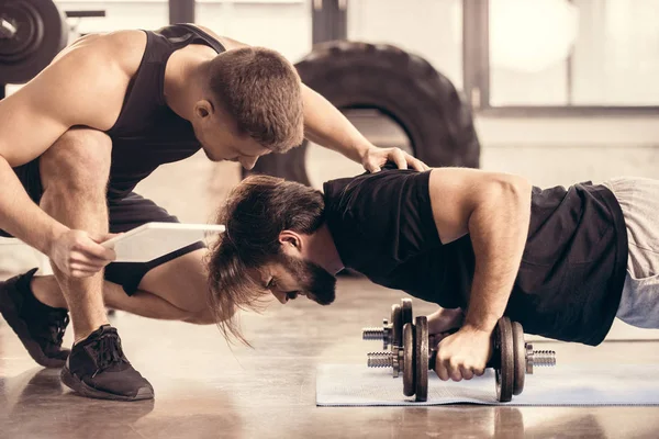 Side view of trainer pushing sportsman doing push ups on dumbbells in gym — Stock Photo