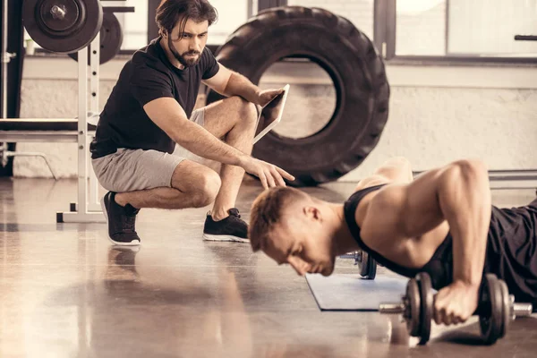 Handsome sportsman doing wide push ups on dumbbells in gym, trainer pointing on something — Stock Photo