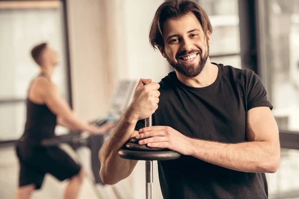 Sportivo sorridente appoggiato alla sbarra di ferro con i dischi in palestra — Foto stock