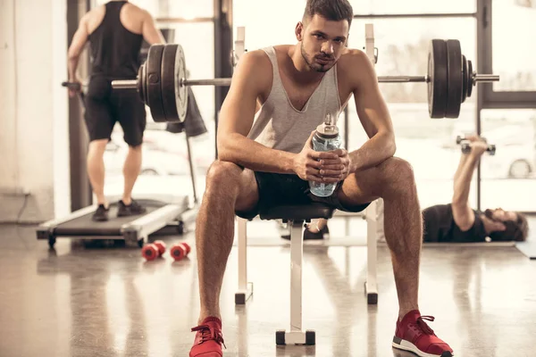 Apuesto deportista sentado con botella de agua en press de banca en el gimnasio - foto de stock