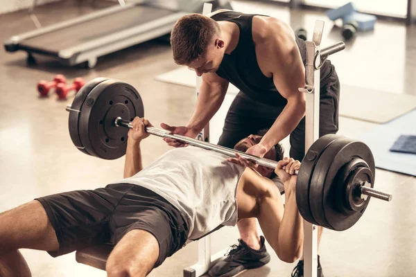 Athletic trainer helping sportsman lifting barbell with heavy weight plates in gym — Stock Photo