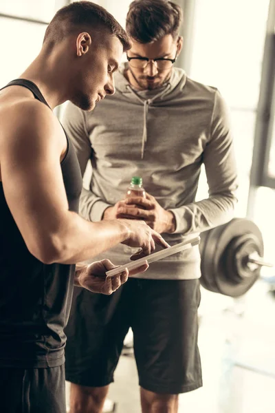 Entrenador guapo que muestra los resultados de hacer ejercicio en la tableta al deportista en el gimnasio - foto de stock
