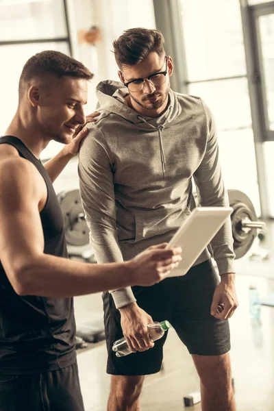 Entrenador guapo que muestra los resultados de hacer ejercicio en la tableta al deportista agotado en el gimnasio - foto de stock