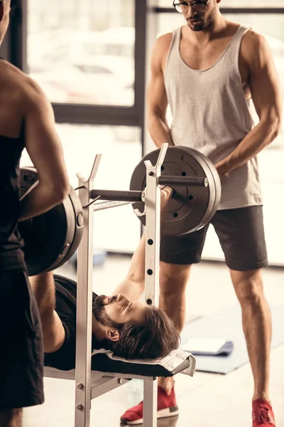 Sportsmen helping friend lifting barbell in gym — Stock Photo