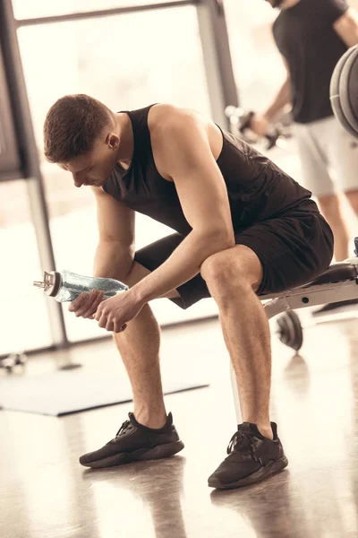 Deportistas guapos descansando y mirando botella de agua en el gimnasio - foto de stock