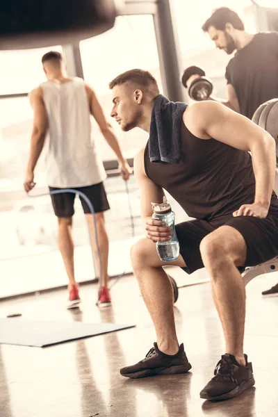 Vue latérale de beaux sportifs se reposant et tenant une bouteille d'eau dans la salle de gym — Photo de stock