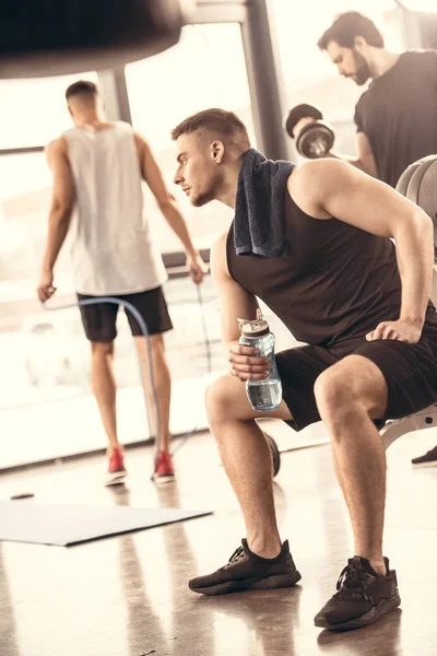 Vista laterale di bei sportivi che riposano e tengono in mano una bottiglia d'acqua in palestra — Foto stock