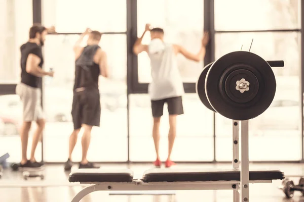 Rear view of sportsmen standing near windows in gym with barbell on foreground — Stock Photo