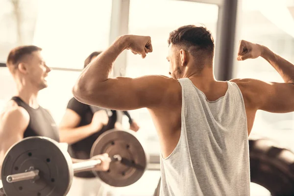 Vista posterior de joven deportista mostrando los músculos a amigos en el gimnasio - foto de stock