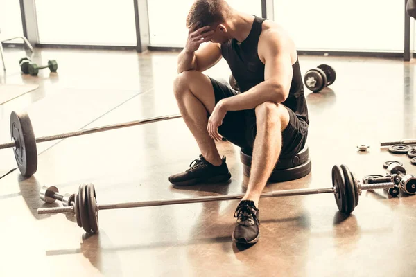 High angle view of upset young sportsman sitting with dumbbells and barbells in gym — Stock Photo