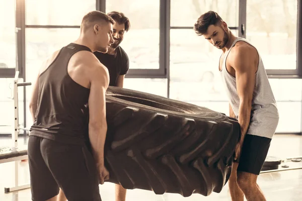 Athletic young sportsmen lifting tyre together in gym — Stock Photo