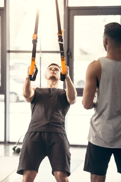 Tiro recortado de hombre deportivo mirando el entrenamiento del hombre muscular con correas de suspensión en el gimnasio - foto de stock