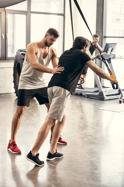 Vista lateral del entrenador ayudando al deportista a hacer ejercicio con correas de suspensión en el gimnasio - foto de stock