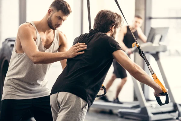 Trainer helping sportsman exercising with suspension straps in gym — Stock Photo