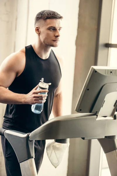 Спортивний молодий чоловік тримає пляшку води і тренується на біговій доріжці — стокове фото