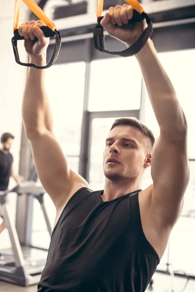 Bel giovane atletico che si allena con cinghie da fitness e distoglie lo sguardo in palestra — Foto stock