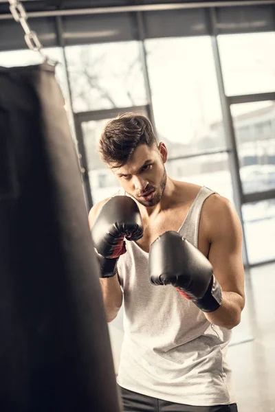 Концентрированный молодой спортсмен в боксёрских перчатках смотрит на боксёрскую грушу в спортзале — стоковое фото