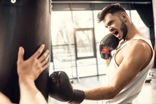 Entrenamiento agresivo joven boxeador con saco de boxeo en el gimnasio - foto de stock