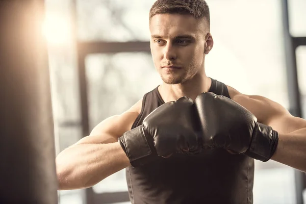Guapo musculoso joven deportista en guantes de boxeo mirando hacia otro lado mientras entrena en el gimnasio - foto de stock