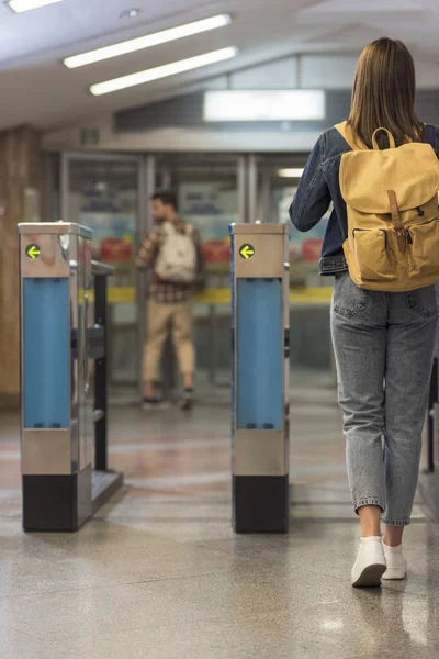 Rear view of female stylish traveler with backpack passing through turnstiles and male tourist behind — Stock Photo