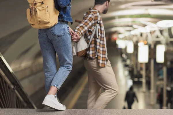 Recortado disparo de un par de turistas con estilo con mochilas cogidas de la mano en la estación de metro - foto de stock