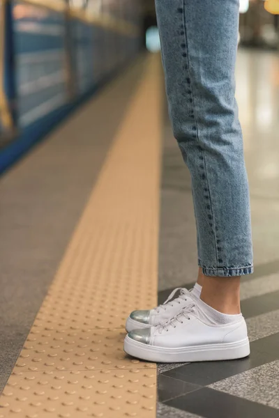 Imagen recortada de piernas femeninas en jeans elegantes y zapatillas de deporte en la estación de metro - foto de stock