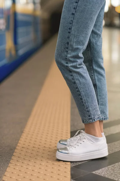 Imagen recortada de piernas femeninas en jeans elegantes y zapatillas de deporte en la estación de metro - foto de stock