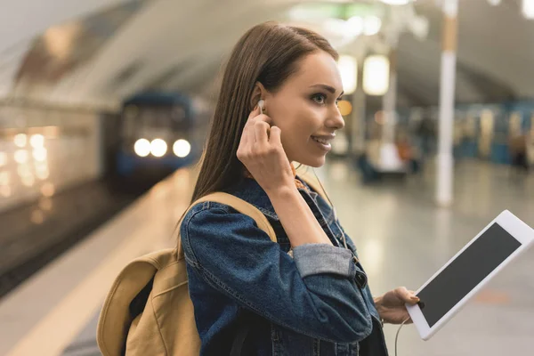 Вид сбоку улыбающейся молодой женщины с наушниками и цифровым планшетом на станции метро — стоковое фото