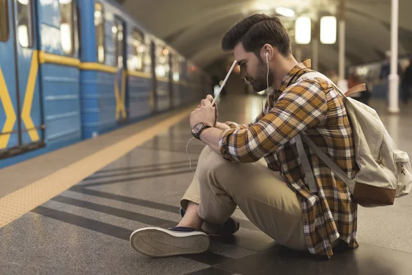 Jeune homme avec tablette numérique et écouteurs assis sur le sol à la station de métro — Photo de stock