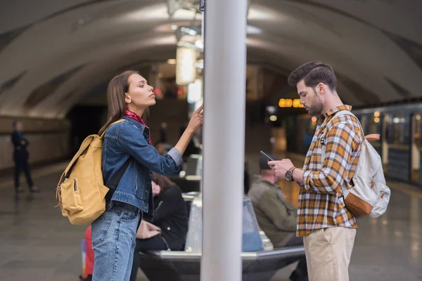 Vista lateral del viajero femenino con mochila mirando el tablero de información y el novio usando el teléfono inteligente en la estación de metro - foto de stock