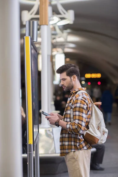 Vista lateral del joven turista masculino con mochila y teléfono inteligente mirando el tablero de información en la estación de metro - foto de stock