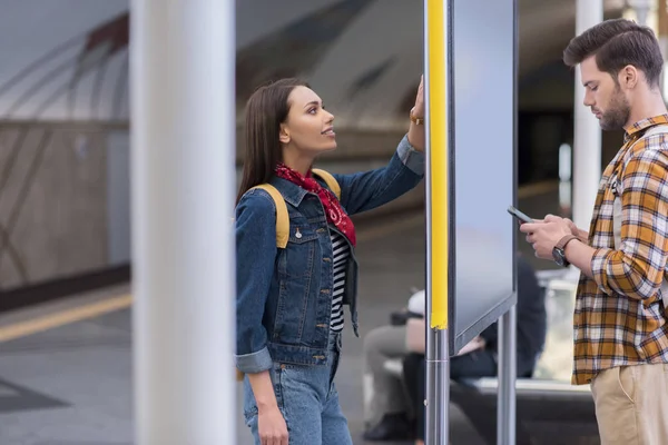 Vista lateral del turista masculino con teléfono inteligente y su novia mirando el tablero de información en la estación de metro - foto de stock
