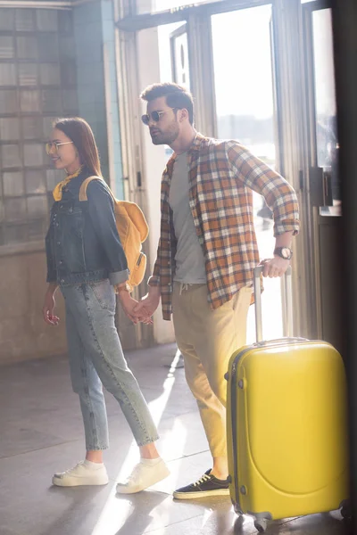 Стильная пара туристов в солнечных очках с рюкзаком и сумкой на колесах в метро — стоковое фото