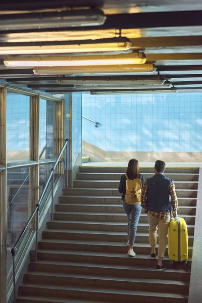 Vista trasera de pareja elegante con mochila y bolsa de ruedas que sube en el metro - foto de stock