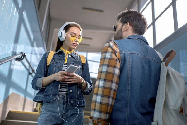 Vista trasera del viajero masculino con mochila y novia con estilo con teléfono inteligente y auriculares en las escaleras del metro - foto de stock